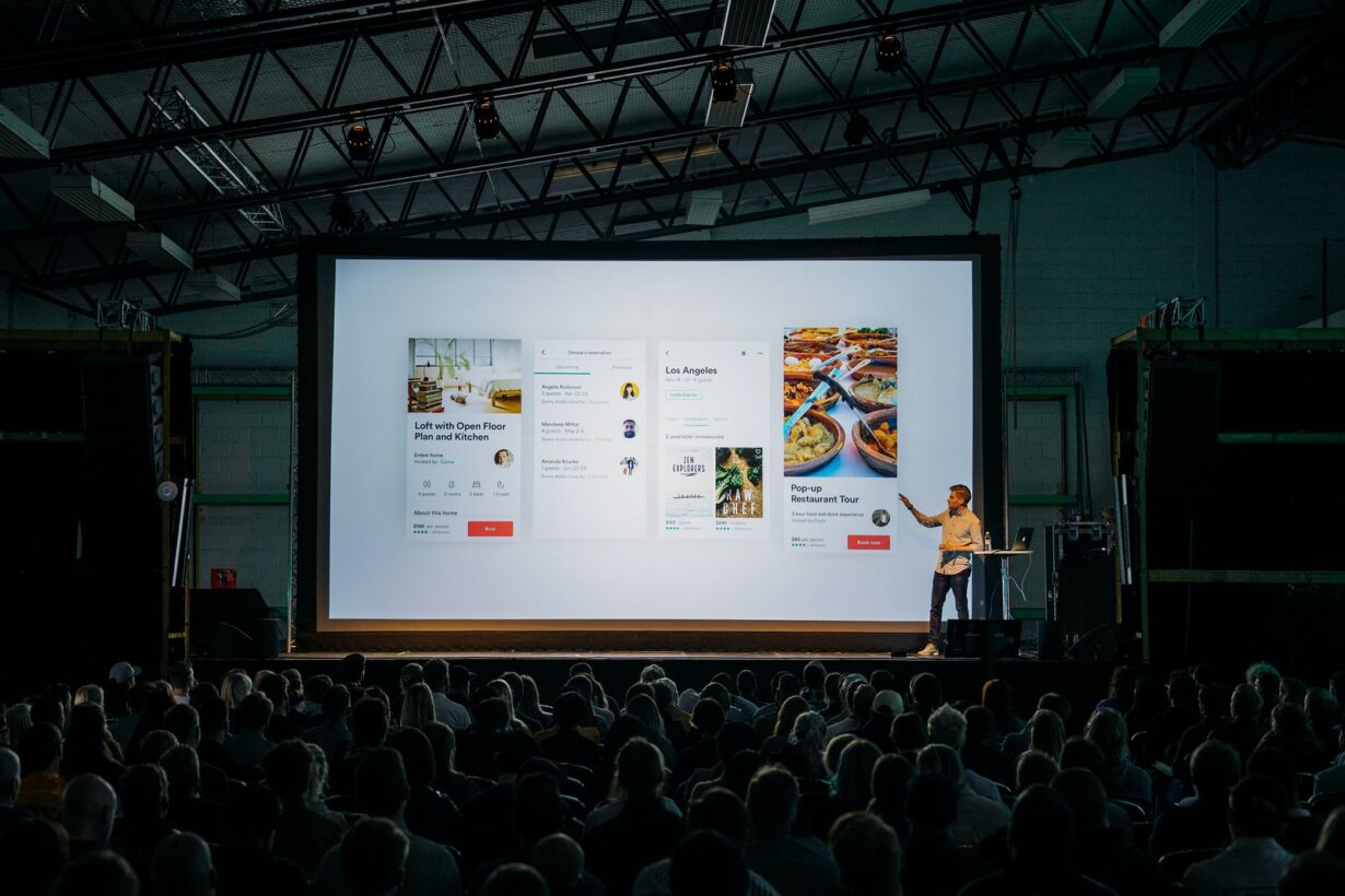 person discussing while standing in front of a large screen in front of people inside dim-lighted room